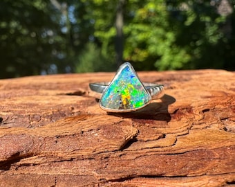 May include: A silver ring with a triangular opal stone. The opal displays a vibrant array of colors, including blue, green, yellow, and orange. The ring is set against a natural wood surface, with a blurred green background.