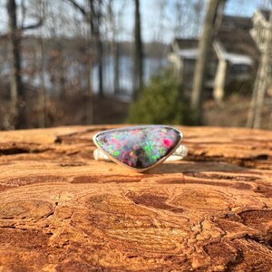 May include: A silver ring with a triangular, multi-colored opal stone. The opal displays a vibrant array of colors including pink, green, and black. The ring is set against a textured, brown wooden surface.