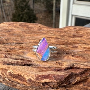 May include: A silver ring with a teardrop-shaped stone. The stone features a blend of purple, blue, and tan colors. The ring is set on a textured, brown wooden surface. The ring's band has a double-band design.