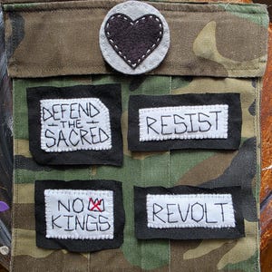 May include: Camouflage fabric pouch with four embroidered patches. The patches read "DEFEND THE SACRED", "RESIST", "NO KINGS", and "REVOLT". A felt heart is sewn on the top flap.