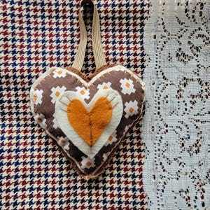 May include: A heart-shaped ornament with a brown and white patterned design, featuring an orange heart in the center. It has a beige loop for hanging. The ornament is placed on a houndstooth pattern fabric.