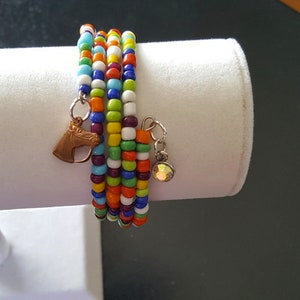May include: A multi-strand wrap bracelet with colorful glass beads in a rainbow of hues. The bracelet features a copper horse head charm and a silver charm with a faceted crystal. The bracelet is displayed on a white cylindrical stand.