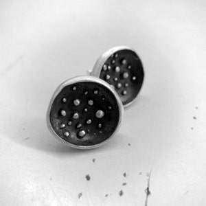 Op de afbeelding: Een paar zilveren oorbel studs met een getextureerd, rond ontwerp. De oorbellen hebben een donkergrijze afwerking en zijn versierd met kleine, verhoogde stippen.