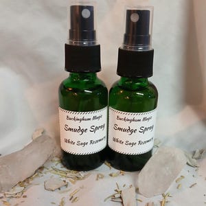May include: Two green glass spray bottles with black spray tops and white labels that read "Buckingham Magic Smudge Spray White Sage Rosemary." The bottles are surrounded by crystals and dried herbs.