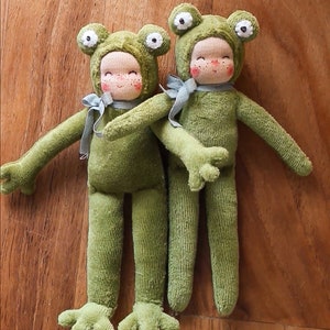 May include: Two handmade green frog dolls with smiling faces, rosy cheeks, and light blue bows. The dolls are wearing green frog costumes with large, white-eyed hoods. The dolls are standing on a wooden surface.