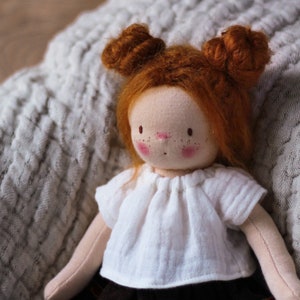 May include: A handmade doll with red hair in two buns, wearing a white shirt and a black skirt. The doll is sitting on a white textured blanket.