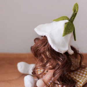Puede incluir: Una muñeca hecha a mano con cabello largo y castaño que lleva un sombrero blanco con forma de flor con hojas de fieltro verde y un vestido de cuadros amarillos y blancos.