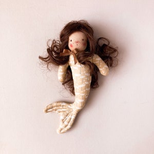 May include: A handmade doll with brown hair, a beige and white patterned dress, and a brown bow. The doll has a painted face with rosy cheeks and a sweet expression.
