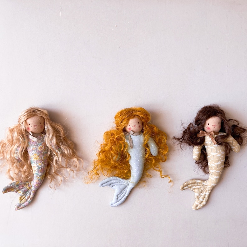 May include: Three handmade fabric mermaid dolls with different colored hair. The dolls have long, curly hair and are wearing different patterned dresses. The dolls are all laying on a white surface.