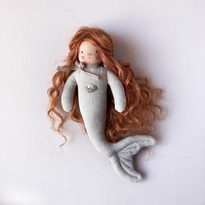 May include: A handmade doll with a light blue mermaid tail and long, curly red hair. The doll has a simple embroidered face and a floral design on the chest.