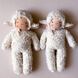 Könnte beinhalten: Zwei weiße Plüsch-Lamm-Puppen mit rosigen Wangen und gestickten Nasen. Die Lämmer tragen weiße flauschige Hüte und haben weiße Schleifenbänder um den Hals.