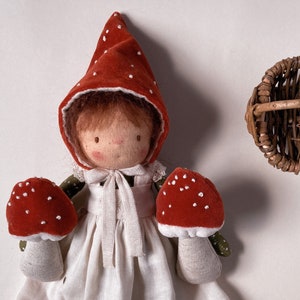 Puede incluir: Una muñeca hecha a mano que lleva un vestido rojo con capucha y sostiene dos juguetes con forma de hongo rojo y blanco. La muñeca tiene el pelo castaño y una cara blanca con rasgos pintados. Una pequeña cesta de mimbre está en el fondo.
