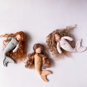 May include: Three handmade fabric mermaid dolls with long curly hair. The mermaids are wearing different colored outfits: light blue, beige, and white.