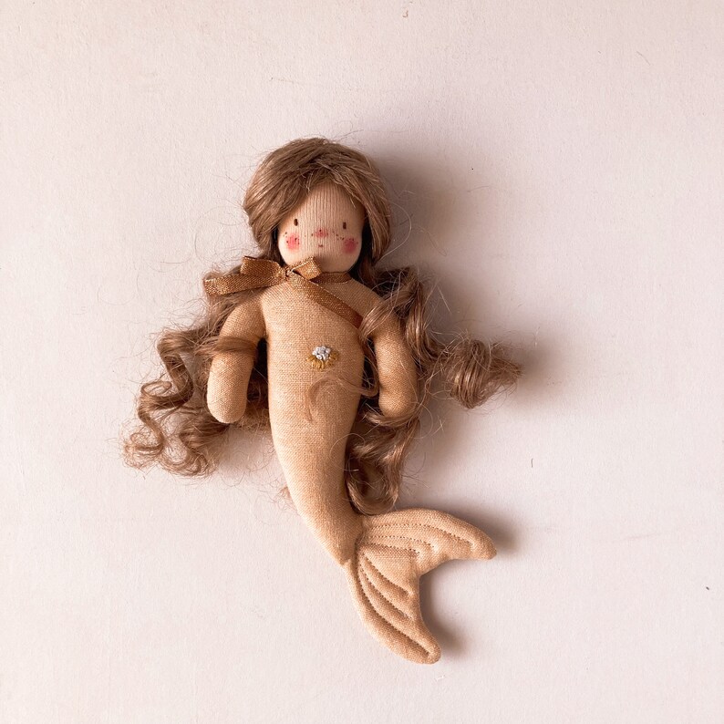 May include: A handmade doll with a brown curly wig, a beige dress, and a tail. The doll has a painted face with rosy cheeks and a small embroidered flower on its chest.