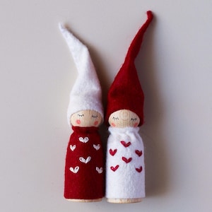 May include: Two felt Christmas gnomes with white and red hats and embroidered hearts. The gnome on the left is wearing a red dress with white hearts and the gnome on the right is wearing a white dress with red hearts.