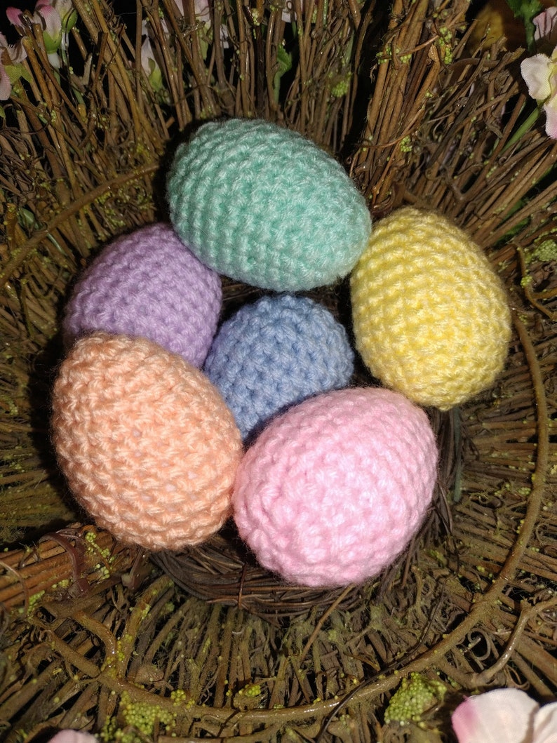 May include: Six crocheted Easter eggs in a nest of twigs. The eggs are in pastel colors: light blue, pink, peach, yellow, lavender, and green.