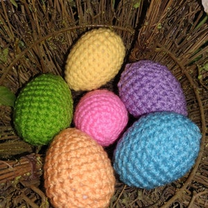 May include: Six crocheted Easter eggs in a nest of twigs. The eggs are yellow, green, purple, pink, orange, and blue.