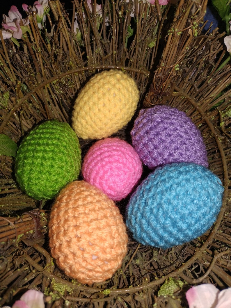 May include: Six crocheted Easter eggs in a nest of twigs. The eggs are yellow, green, purple, pink, orange, and blue.