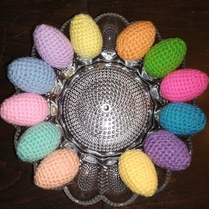 May include: A collection of crocheted Easter eggs in various colors, including pink, blue, green, yellow, and orange, arranged in a circle on a clear glass plate with a textured surface.