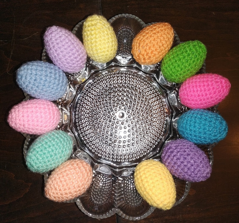 May include: A collection of crocheted Easter eggs in various colors, including pink, blue, green, yellow, and orange, arranged in a circle on a clear glass plate with a textured surface.