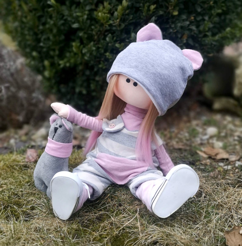 May include: A handmade doll with pink and grey striped overalls, a grey hat with pink ears, and blonde hair with pink streaks. The doll is sitting on a bed of green grass and holding a grey stuffed animal.