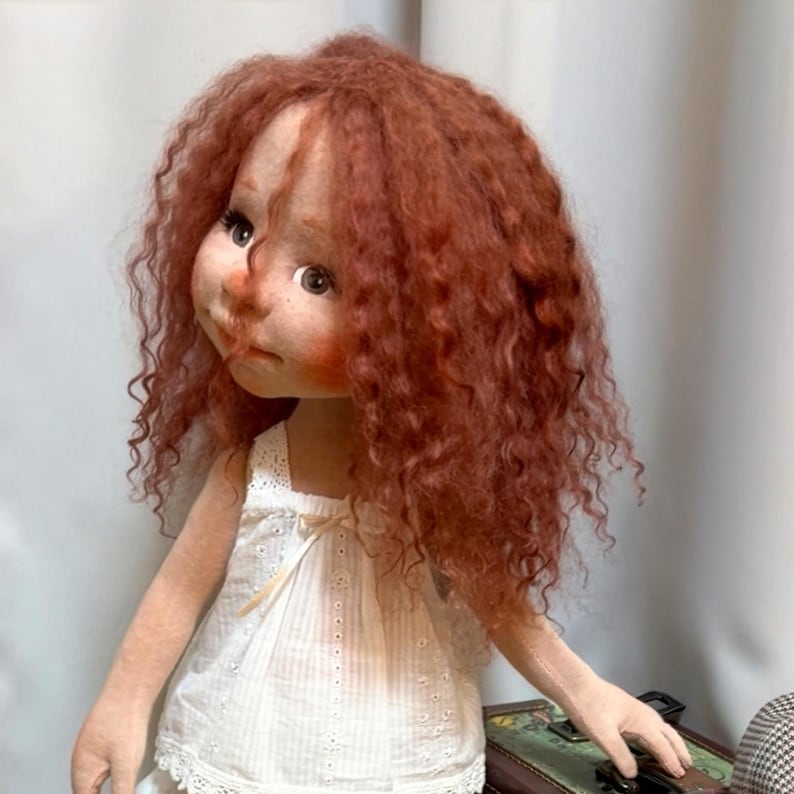 May include: A handmade doll with a whimsical design. The doll has red curly hair, rosy cheeks, and is wearing a white eyelet dress. The doll is a unique piece of art, perfect for collectors or as a decorative item.
