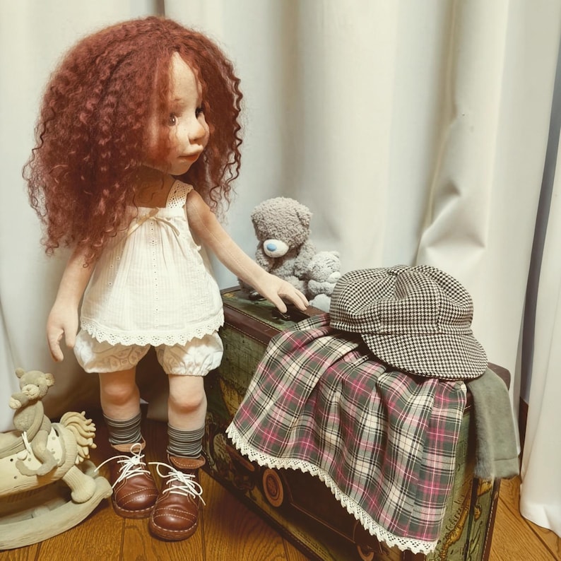 May include: A handmade doll with red curly hair, wearing a white dress, bloomers, and brown shoes. A plaid skirt, a houndstooth cap, and a teddy bear are on a vintage trunk. A rocking horse is in the foreground.