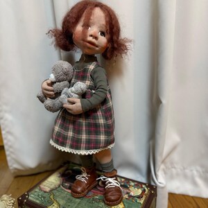 May include: A handmade doll with red hair in pigtails, wearing a green long-sleeved shirt and a plaid dress with a white lace trim. The doll is holding two gray teddy bears and wearing brown leather shoes with white laces. The doll is standing on a vintage map-covered chest.