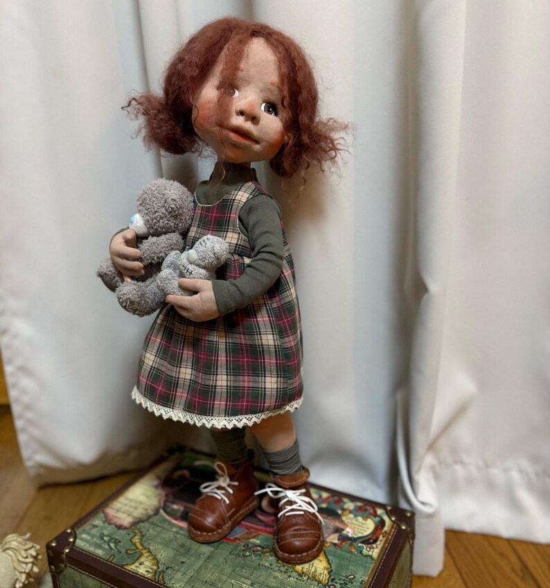 May include: A handmade doll with red hair in pigtails, wearing a green long-sleeved shirt and a plaid dress with a white lace trim. The doll is holding two gray teddy bears and wearing brown leather shoes with white laces. The doll is standing on a vintage map-covered chest.