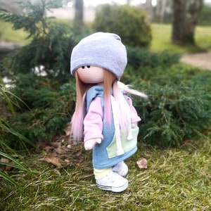 May include: A handmade doll with long pink and brown hair, wearing a blue and green hoodie, a grey beanie, and white sneakers. The doll is standing on green grass.