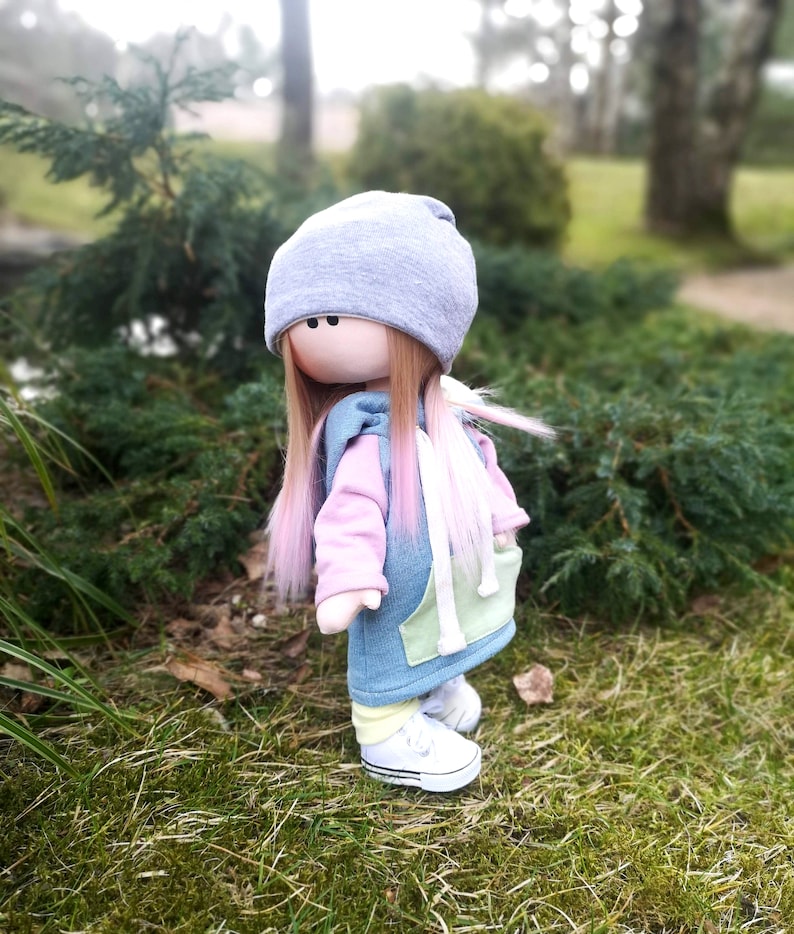 May include: A handmade doll with long pink and brown hair, wearing a blue and green hoodie, a grey beanie, and white sneakers. The doll is standing on green grass.