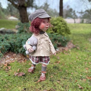 May include: A handmade doll dressed in a tan jacket, plaid skirt, and brown shoes. The doll has red curly hair, a checkered cap, and is holding a gray teddy bear. The doll is standing on green grass.
