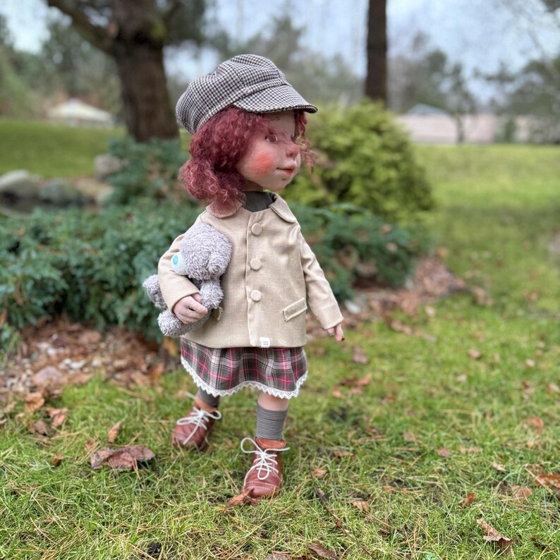 May include: A handmade doll dressed in a tan jacket, plaid skirt, and brown shoes. The doll has red curly hair, a checkered cap, and is holding a gray teddy bear. The doll is standing on green grass.