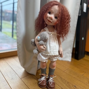 May include: A handmade doll with red curly hair, wearing a white dress, shorts, and brown shoes. The doll is holding a small gray teddy bear. The doll is standing on a wooden floor.