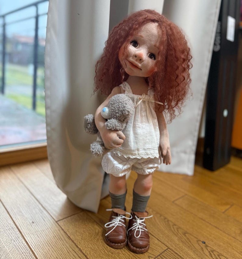 May include: A handmade doll with red curly hair, wearing a white dress, shorts, and brown shoes. The doll is holding a small gray teddy bear. The doll is standing on a wooden floor.