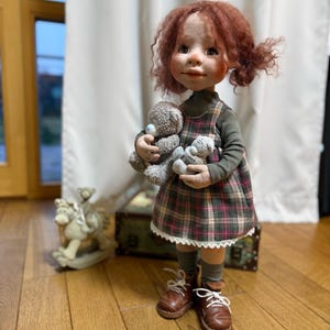 May include: A handcrafted doll with red curly hair, wearing a plaid dress and holding two gray teddy bears. The doll is dressed in a green long-sleeved shirt, plaid dress, and brown boots. A rocking horse is in the background.