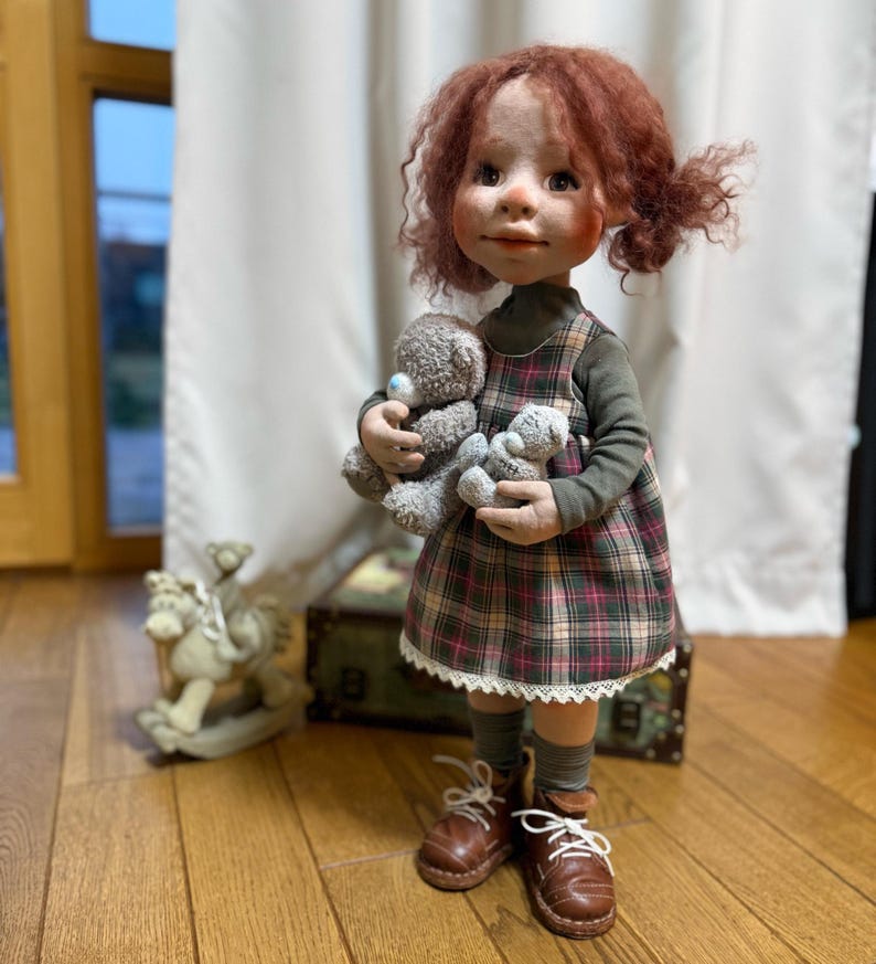 May include: A handcrafted doll with red curly hair, wearing a plaid dress and holding two gray teddy bears. The doll is dressed in a green long-sleeved shirt, plaid dress, and brown boots. A rocking horse is in the background.