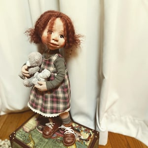 Może przedstawiać: Ręcznie robiona lalka z rudymi kręconymi włosami, ubrana w sukienkę w kratę na bluzce z długim rękawem i brązowe buty. Lalka trzyma dwa szare misie. Stoi na walizce w stylu vintage.