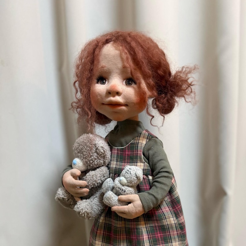 May include: A handcrafted doll with red curly hair and a plaid dress, holding two small gray teddy bears. The doll has a detailed face with rosy cheeks and is wearing a green long-sleeved shirt underneath the dress.