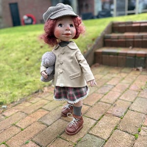 May include: A handcrafted doll with red curly hair, wearing a tan coat, plaid skirt, and brown boots. The doll is holding a gray stuffed animal and a gray and white checkered cap. The doll is standing on a brick walkway.