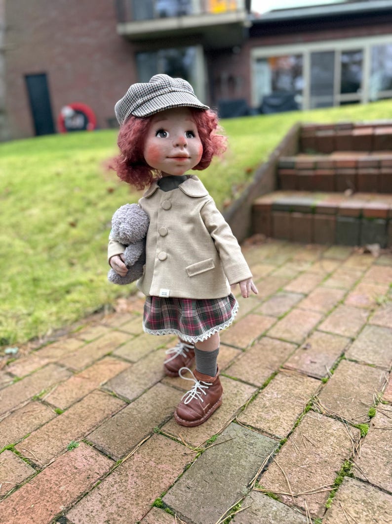 May include: A handcrafted doll with red curly hair, wearing a tan coat, plaid skirt, and brown boots. The doll is holding a gray stuffed animal and a gray and white checkered cap. The doll is standing on a brick walkway.