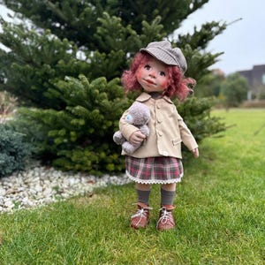 May include: A handmade doll with red curly hair, wearing a tan jacket, plaid skirt, and brown shoes. The doll is holding a gray teddy bear and a gray and white hat. The doll is standing on green grass.