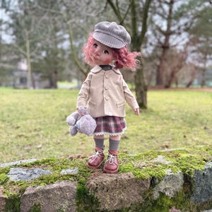 May include: A doll dressed in a tan jacket, plaid skirt, and brown boots, holding a gray stuffed animal. The doll has pink hair and a gray plaid cap. It is standing on a moss-covered stone wall with a blurred green background.
