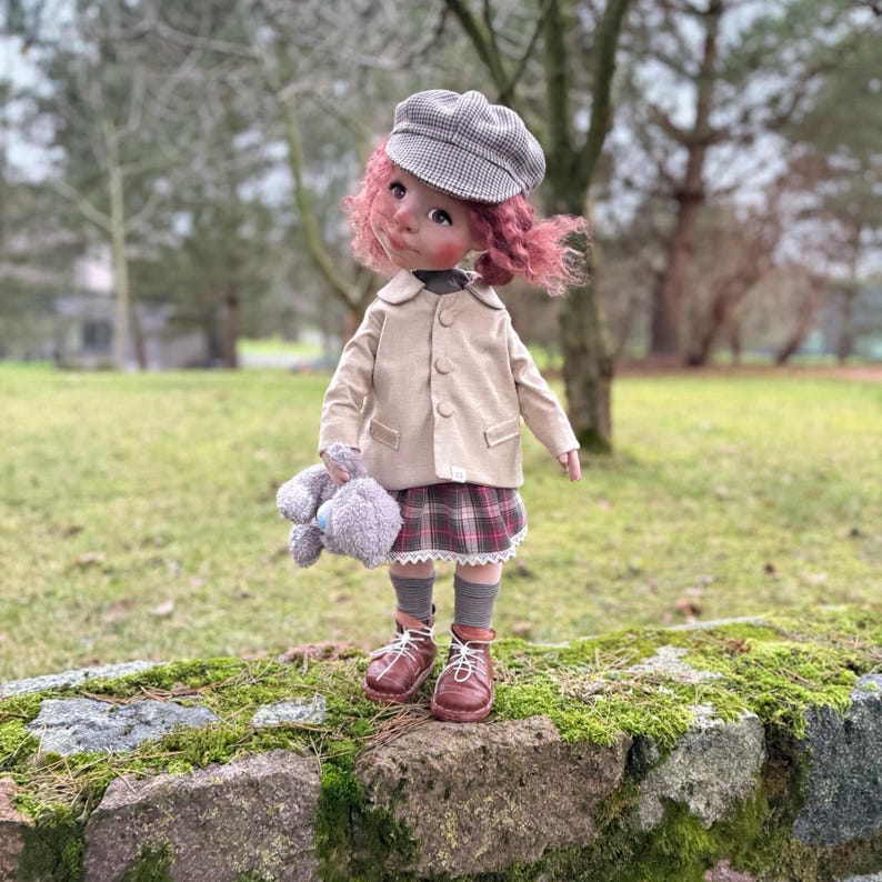 May include: A doll dressed in a tan jacket, plaid skirt, and brown boots, holding a gray stuffed animal. The doll has pink hair and a gray plaid cap. It is standing on a moss-covered stone wall with a blurred green background.