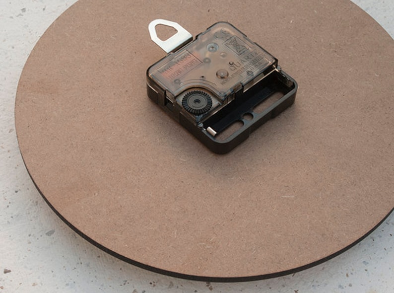 May include: A close-up of a brown wooden clock face with a black clock mechanism attached to the back. The clock mechanism has a clear plastic cover and a silver hook.