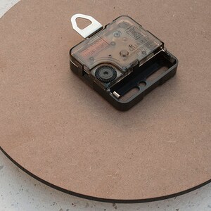 May include: A close-up of a brown circular clock face with a black clock mechanism attached to the back. The clock mechanism has a clear plastic cover and a silver hook.