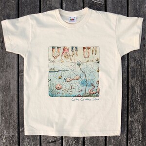 May include: A white t-shirt with a cartoon illustration of children dangling upside down over a sea scene with fish, crabs, and seaweed. The text "Crazy Crabbing, Devon" is printed below the illustration.