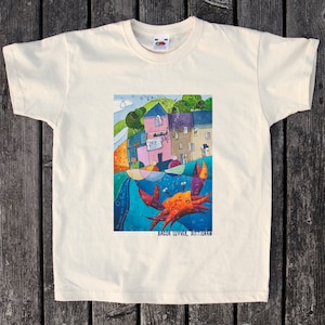 Puede incluir: Una camiseta blanca con una ilustración colorida de una ciudad con un cangrejo en el agua. El texto "BACON LOVER. DITISHAM" está impreso debajo de la imagen.