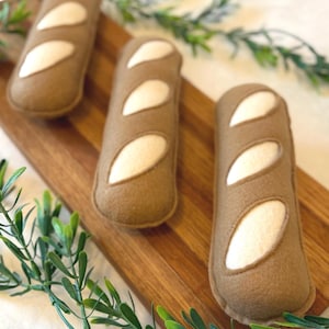 May include: Three handcrafted felt baguette toys in a light brown colour with cream-coloured diagonal slash details on the top surface. These soft, plush bread-shaped items are arranged in a row on a rectangular wooden board with green foliage.
