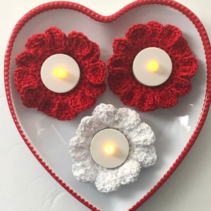 Puede incluir: Tres portavelas de ganchillo en un plato con forma de corazón. Dos son rojos y uno es blanco. Cada portavelas tiene una pequeña vela de té dentro.